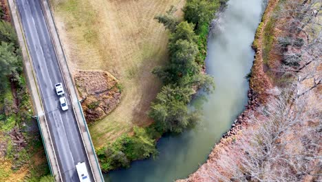 drone footage tracks vehicles driving across a two-lane bridge spanning a winding river in a rural, grassy landscape with scattered trees and soft daylight