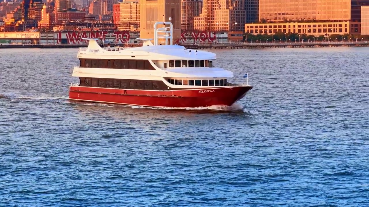 New York, USA, 1. juli 2026: Atlantica turbåd sejler langs Manhattans havnefront. Den rød-hvide Atlantica-færge sejler på floden forbi oplyste moler og skyskrabere ved solnedgang.