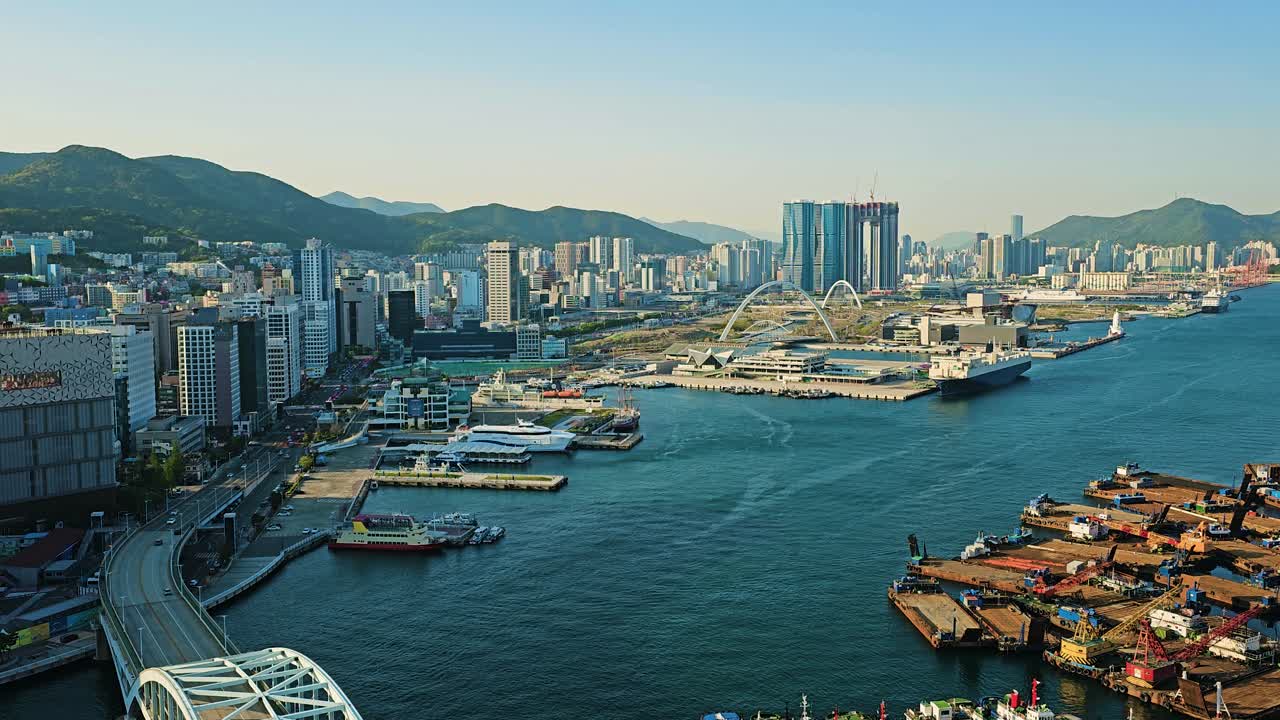 Aerial view of Busan port with cargo ships, cranes, city skyline, and iconic modern buildings under blue sky