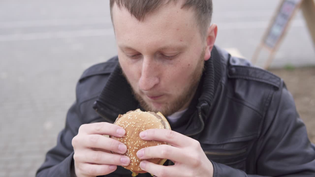 joven con barba en el café de la calle mordiendo sabrosa hamburguesa grande con queso. papas fritas en el plato. filmado en 4k