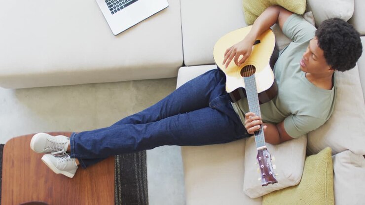 Uomo afroamericano suona la chitarra, canta e usa il laptop a casa.