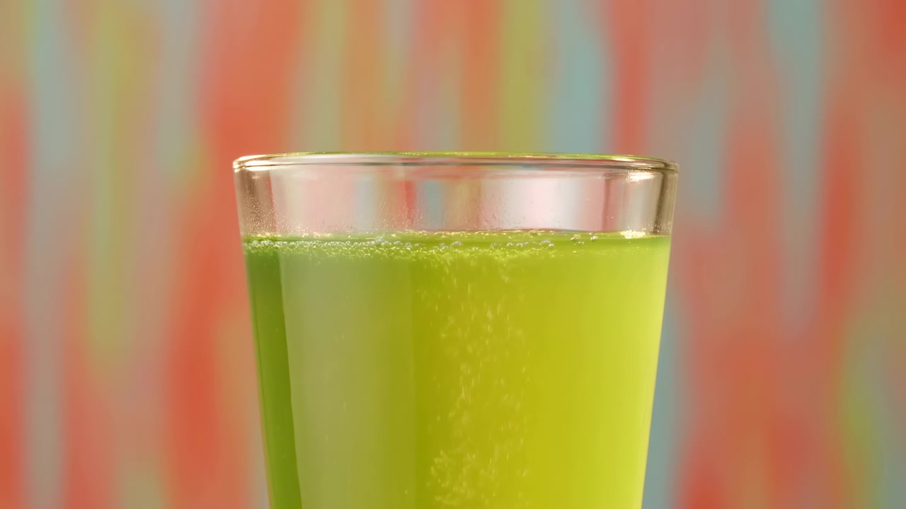 Succo verde vibrante in un bicchiere, primo piano con sfondo sfocato e colorato, bollicine frizzanti lungo il bordo, illuminato in studio.