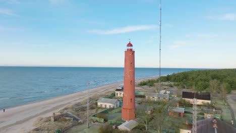 luftaufnahme des rot gefärbten leuchtturms von akmenrags, ostseeküste, lettland, weißer sandstrand, ruhiges meer, sonniger tag mit wolken, breite drohnenaufnahme, die sich vorwärts bewegt, nach unten geneigt