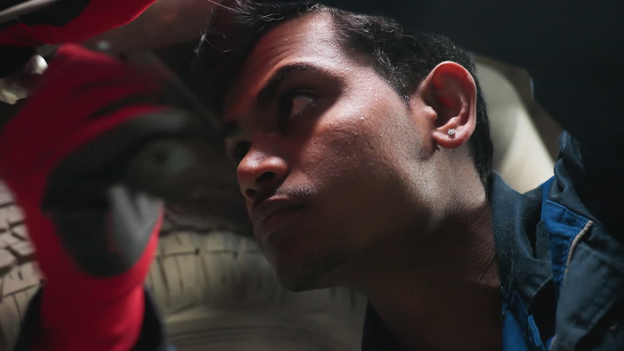 자동차 에서 일하는 기계공, 집중된 표정으로 볼트를 풀고, 그를 위해 빛을 들고 있는 누군가의 부분적인 시각, 그의 얼굴에 반사되는 빛, 장갑과 파란 유니폼을 입고