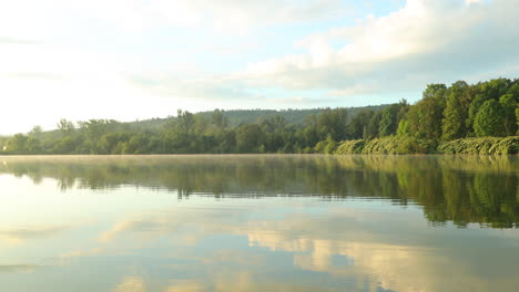 view of the calm water level in the lake where morning mist rushes over the water with the rising sun reflected on the surface and after the calm surface of the lake a fish emerges in the distance