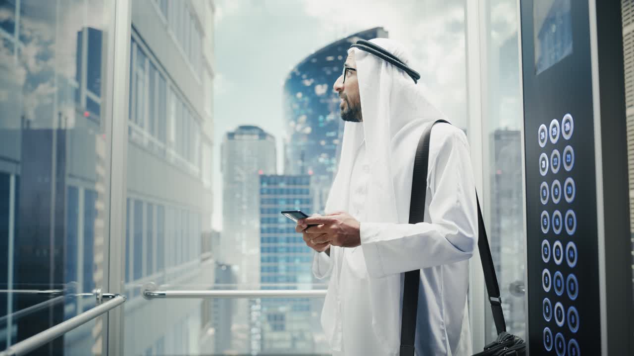 un hombre de negocios musulmán exitoso en el tradicional kandura blanco montando un ascensor de vidrio a la oficina en un centro de negocios moderno. hombre usando un teléfono inteligente. saudi, emirati, concepto de hombre de negocios árabe.