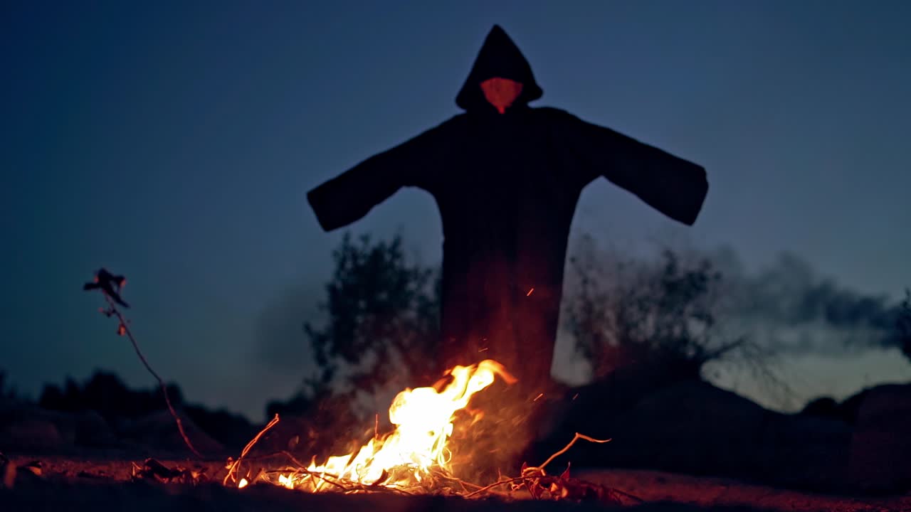 Scary witch stands by the fire at night. Black figure in cloak with hood walking around the fire with outstretched arms. Halloween outdoors.