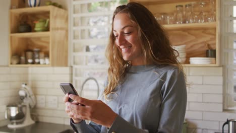 Happy-caucasian-woman-standing-in-sunny-cottage-kitchen-using-smartphone-and-smiling