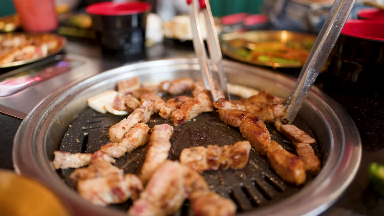 Hand uses metal tongs to flip pork belly on tabletop Korean barbecue grill, shallow focus