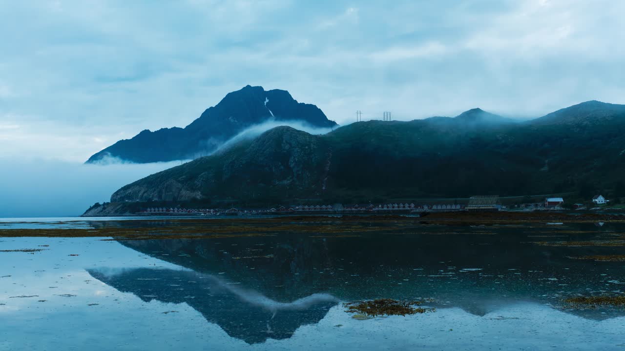 Norwegian Mountains, sea fog and Rorbuer facilities (Traditional fishermen's huts) and their reflections in the water during low tide. Wide angle Time Lapse. Lofoten Islands, Northern Norway