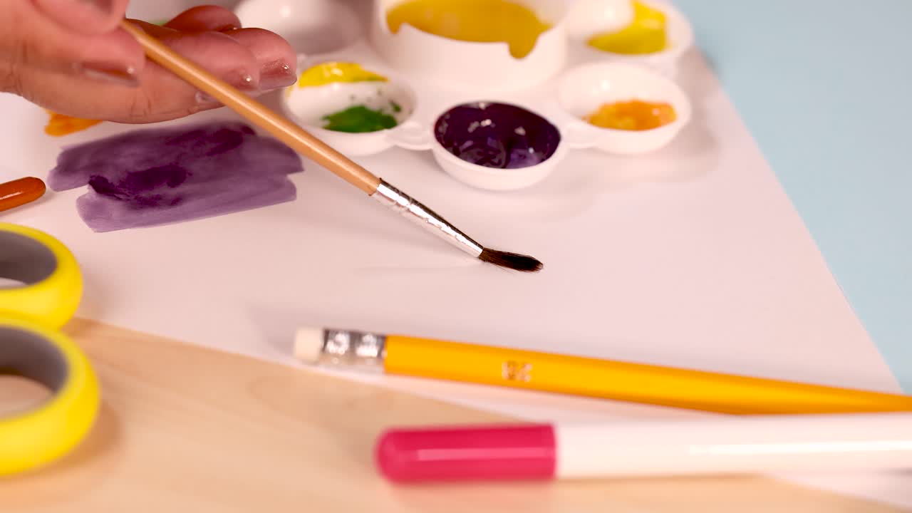 Artist hand uses brush and palette to paint colorful strokes on white paper, bright lighting