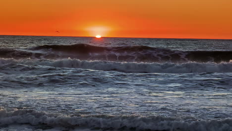 Toma-De-Tiempo-De-Una-Majestuosa-Puesta-De-Sol-A-Lo-Largo-Del-Cielo-Naranja-Sobre-El-Horizonte-Con-Olas-Rompiendo-A-Lo-Largo-De-La-Playa-De-Arena-Durante-La-Noche