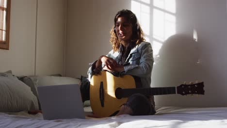 Relaxed-mixed-race-woman-in-headphones-sitting-with-guitar-using-laptop-in-sunny-cottage-bedroom