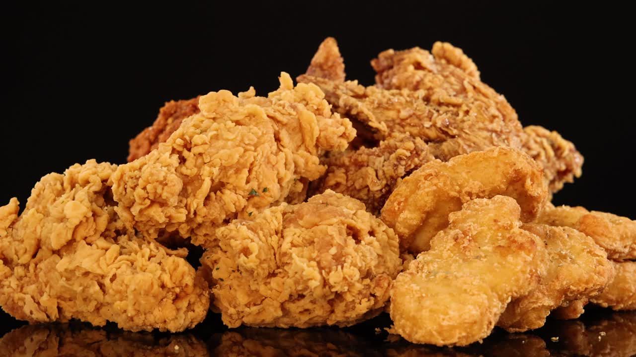 Golden fried chicken wings, drumsticks, and nuggets rotate on a reflective surface under dramatic studio lighting, highlighting crunchy textures and appetizing details