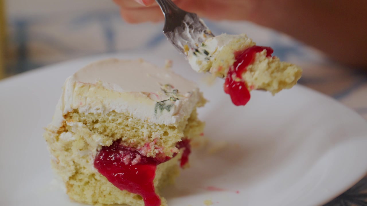 Hand cuts slice of cherry cake on plate. Creamy frosting drips as fork lifts moist crumb. Sweet berry filling oozes from layered cake. Soft focus captures indulgence in dessert moment
