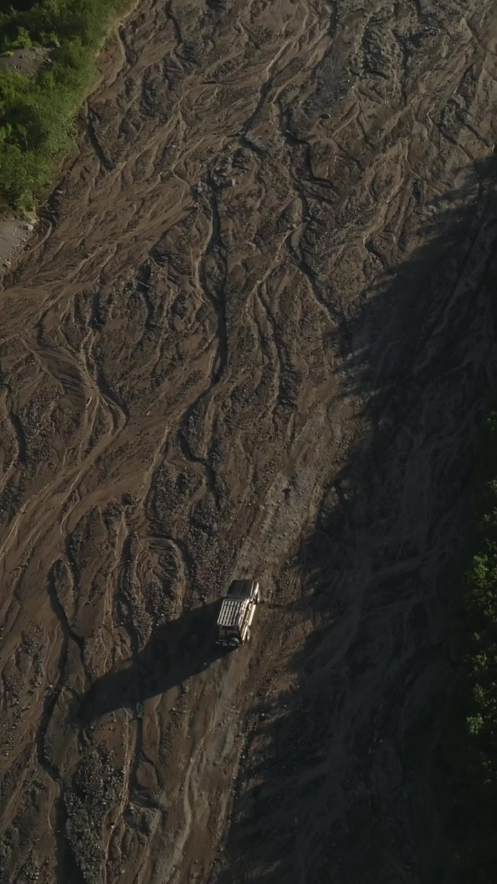 High aerial drone shot of a lone truck following the dry Sukhaya Riverbed track toward Avachinsky Volcano in Kamchatka Krai, Russia, revealing a rugged green mountain valley in summer light
