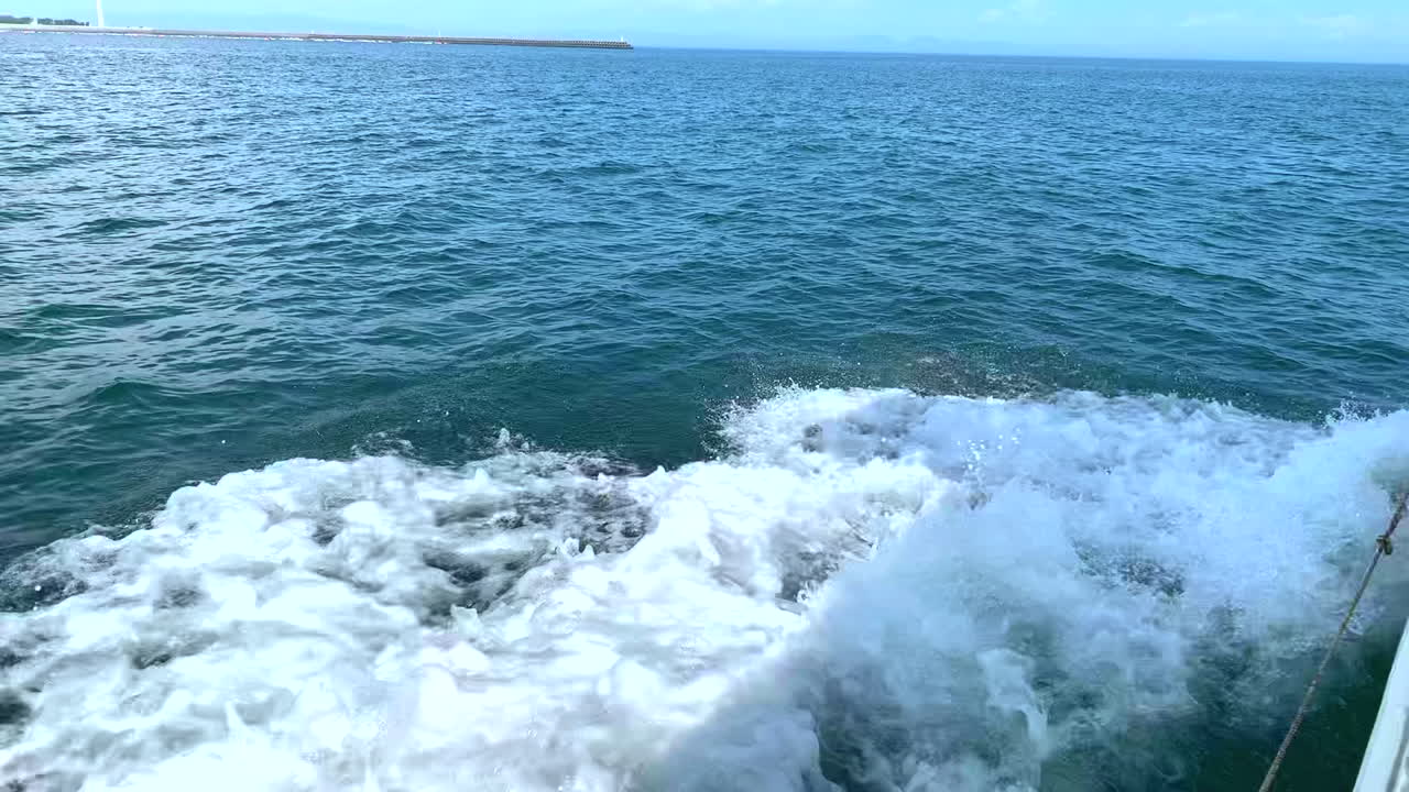 Motor boat sails in the ocean off the coast at full speed in Amakusa Dolphin while watching the trail of white foam leaving and a wind turbine on the background beach shore in a sunny day