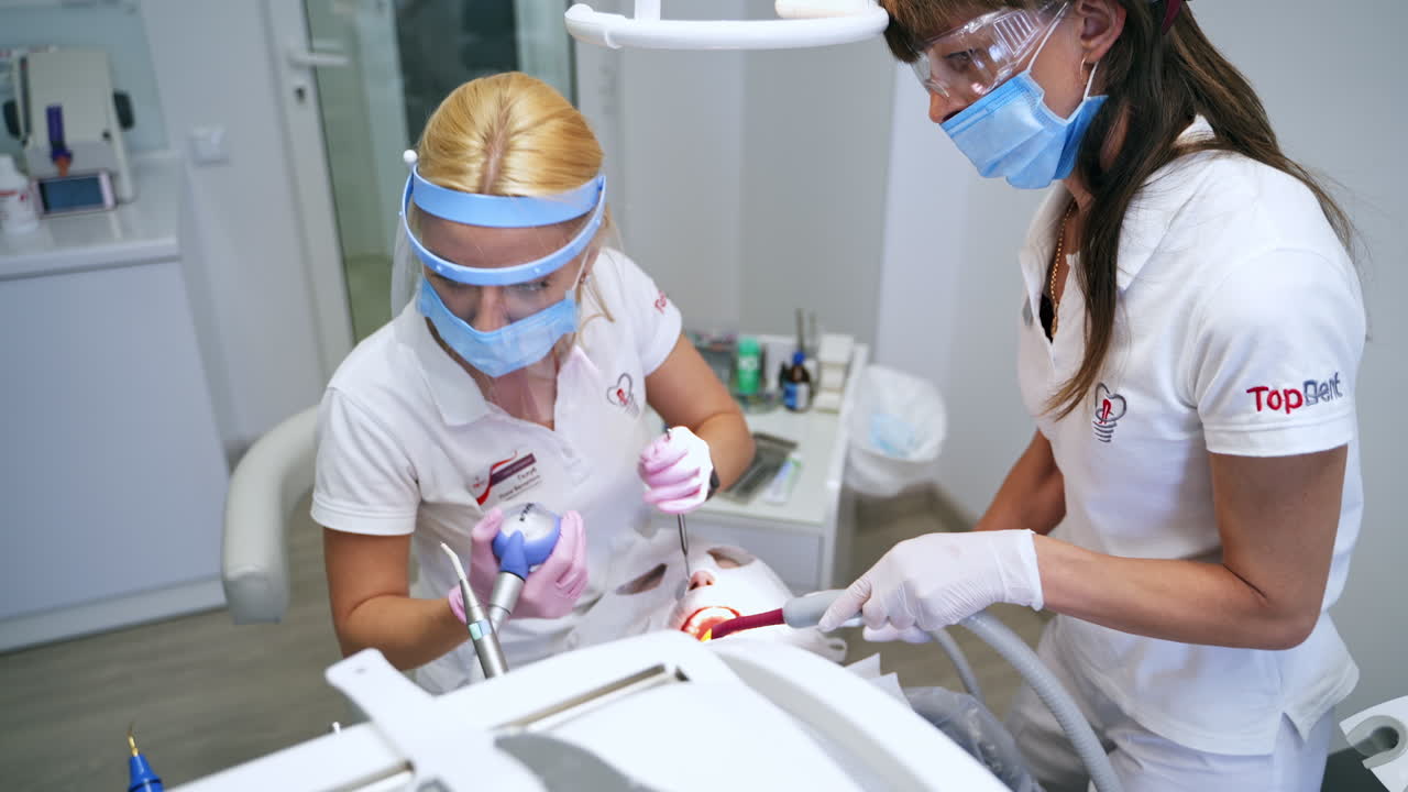 Mid-aged female dentist wearing protective shield bent over the patient. Doctor changes the device of drill machine. Top view.