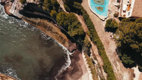 Vista-Aérea-De-Las-Olas-Rompiendo-Contra-La-Orilla-De-Una-Casa-Con-Escaleras-Al-Mar-En-Ametlla-De-Mar,-Tarragona,-España