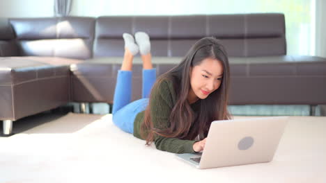 Junge-Glückliche-Frau,-Die-Auf-Dem-Boden-Des-Wohnzimmers-Liegt-Und-Mit-Einem-Lächeln-Auf-Ihrem-Gesicht-Auf-Einem-Laptop-Tippt