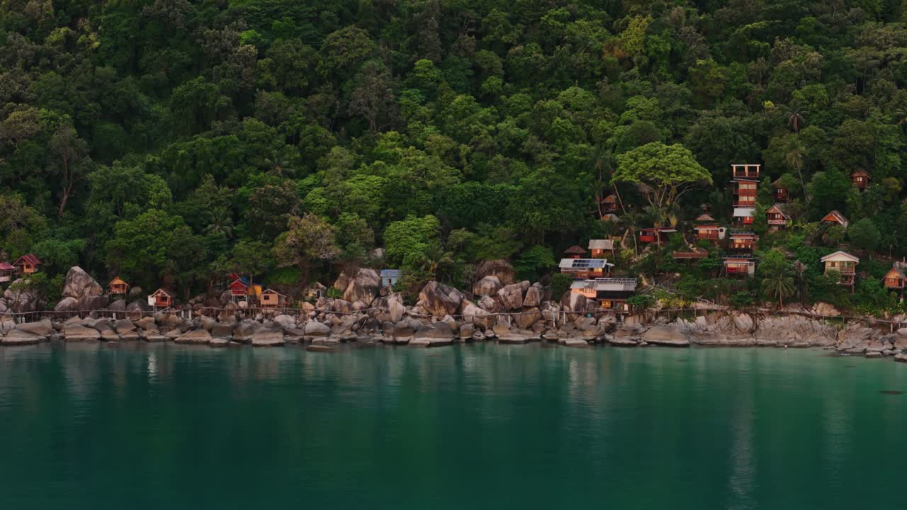Aerial footage of a rocky tropical coastline with lush jungle and seaside houses in Thailand. Calm turquoise sea, green forest, and scenic shoreline. Perfect for travel, nature, and lifestyle content.