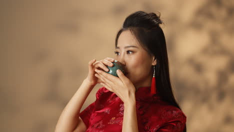 카메라 앞에서 유쾌하게 웃고 컵에서 차를 마시는 붉은 전통 옷을 입은 젊은 아시아 여성의 클로즈업 보기