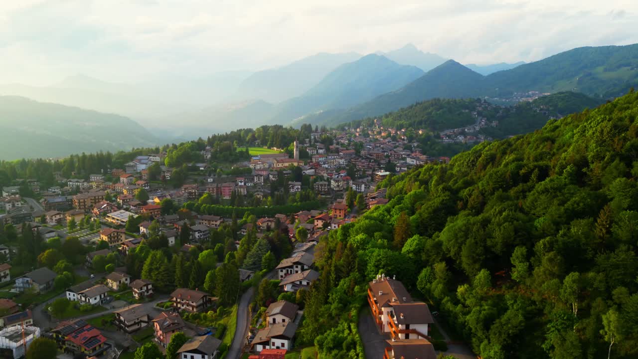 Drone shot of mountain town homes surrounded by forested hills and winding roads under hazy sunlight. Shot at Selvino, Italy (Selvino, Italia)