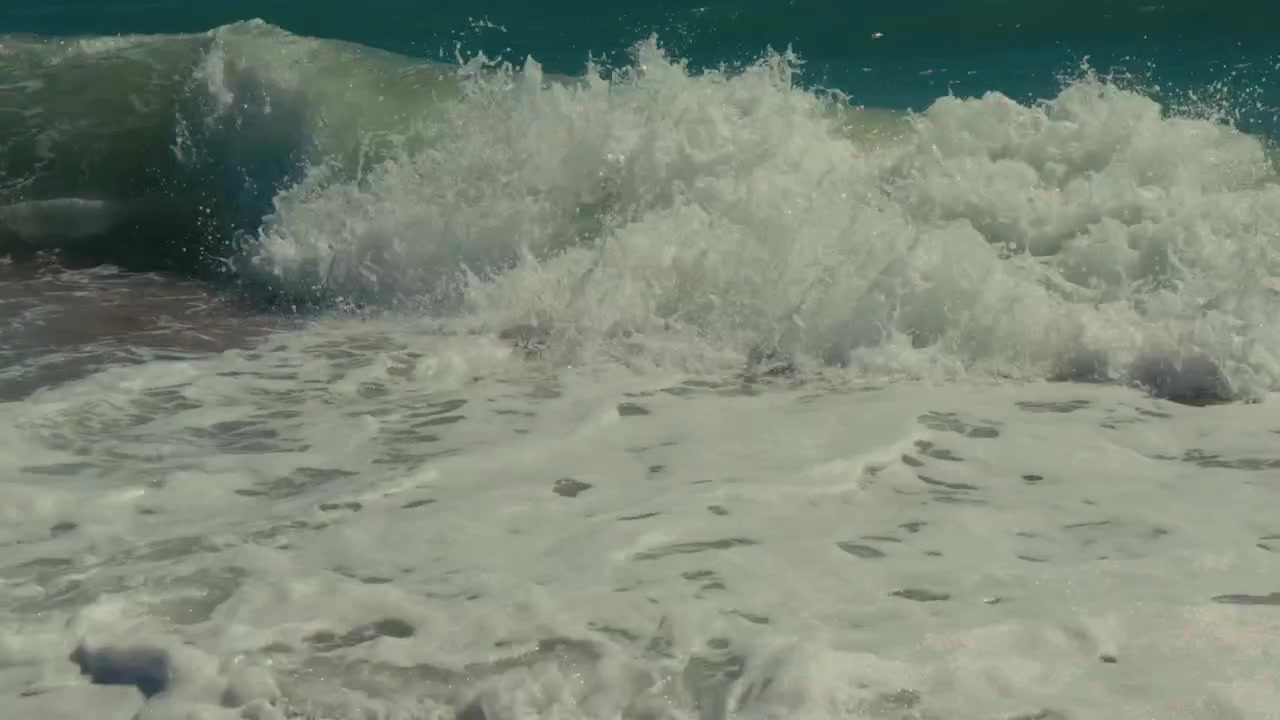 Wave breaking strongly over pebble shoreline, creating white foam and splashing water along coastal beach under bright natural light in Brighton, England.