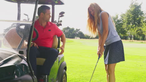 man in golf buggy giving advice to woman as she practices with golf club