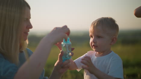 primer plano de una mujer con un vestido azul sosteniendo una botella de espuma en un campo cubierto de hierba mientras un niño con una camiseta blanca observa con entusiasmo, listo para soplar burbujas en el aire