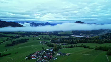 Aerial-drone-shot-flying-high-over-village-houses-with-green-farmlands-surrounded-by-mountain-range-on-a-cloudy-day