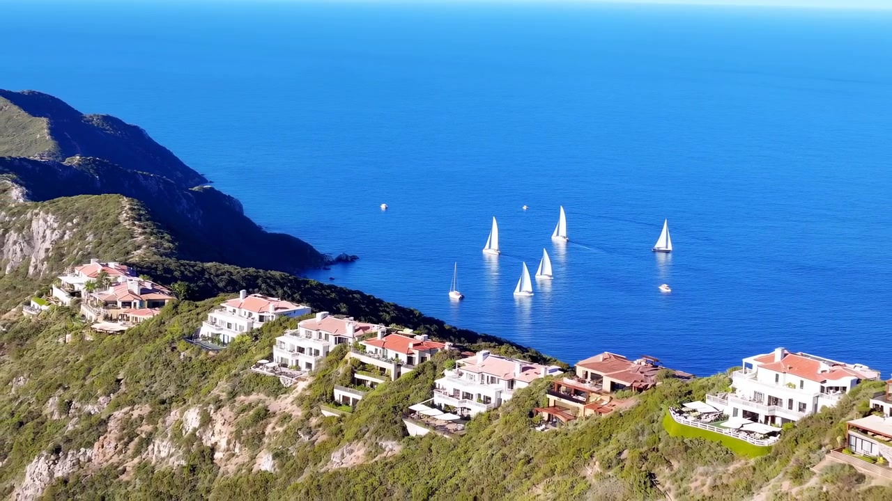 Vista elevada de uma vila costeira com vilas brancas, telhados de telha vermelha, costa rochosa, água azul calma, veleiros à deriva e paisagem litorânea ensolarada sob a luz tranquila do dia, com elementos de costa, mar e oceano