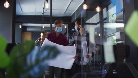 Diverse-male-colleagues-face-masks-discussing-over-strategy-plan-at-modern-office