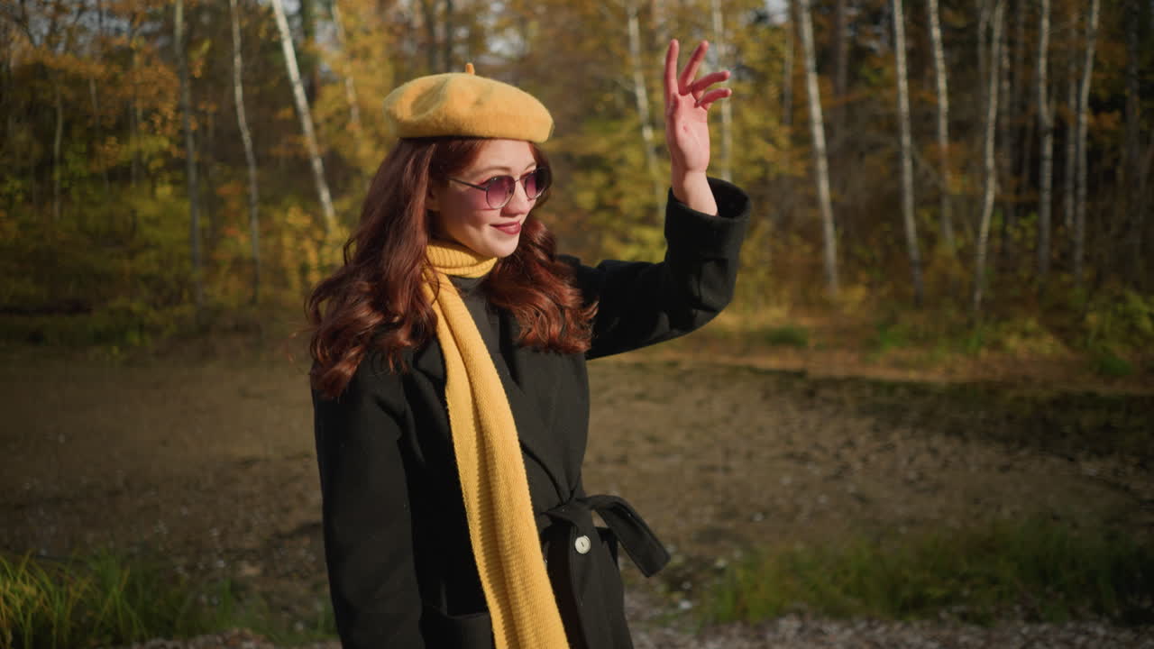 dama con boina amarilla y silenciador camina por el camino del bosque, protegiendo los rayos del sol con su mano, la luz del sol crea un brillo cálido a su alrededor mientras disfruta del pacífico paisaje de otoño
