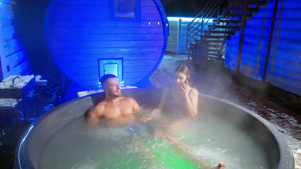 Happy Caucasian people are relaxing in the big hot tub outdoors in winter. Man is enjoying his time while woman is speaking on the phone.