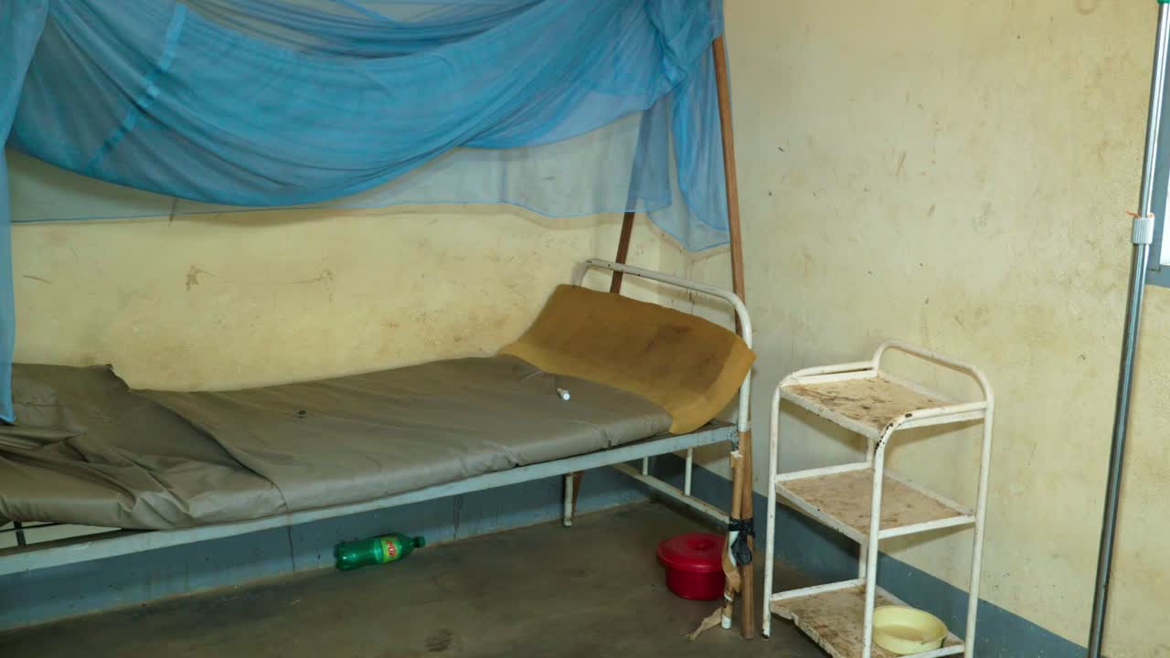 Letto vuoto con zanzariera blu e scaffali arrugginiti in un reparto ospedaliero mal tenuto in Camerun. Inquadratura statica grandangolare