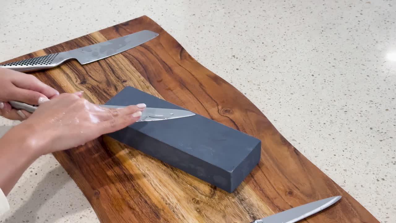 Hands skillfully sharpen a chef’s knife on a whetstone atop a wooden cutting board, under bright, even lighting in a contemporary kitchen setting
