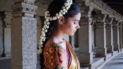 indian woman wearing sari, jasmine adorned braids, performing devotional prayer inside ancient temple with ornate architectural columns surrounding her