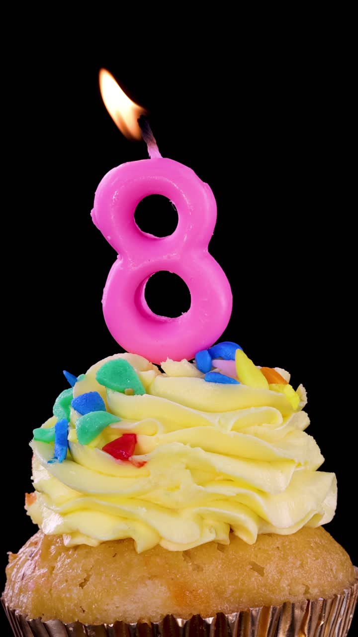 Una candela rosa a forma di numero otto su un cupcake decorato viene spenta, con la fiamma che si estingue e il fumo che sale, su uno sfondo nero.