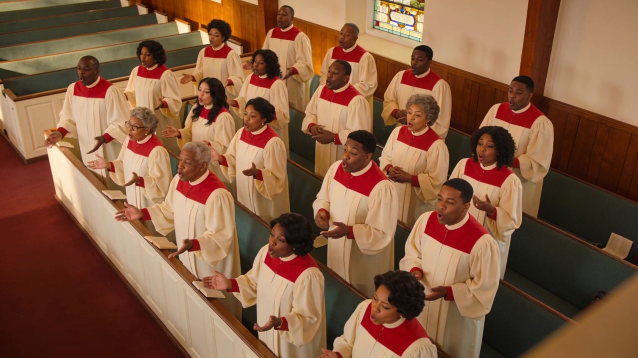 Singing church choir in cream robes with red yokes raising hands during hymn, stained-glass visible