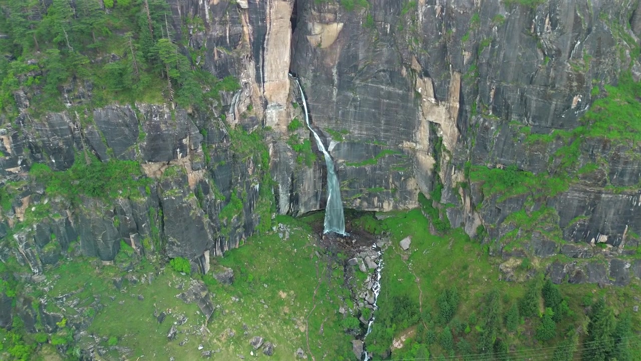 Ripresa aerea con drone in avvicinamento di una stretta cascata che scende da una massiccia scogliera di roccia nera a picco, circondata da erba verde lussureggiante e pini a Manali, Himachal Pradesh, India