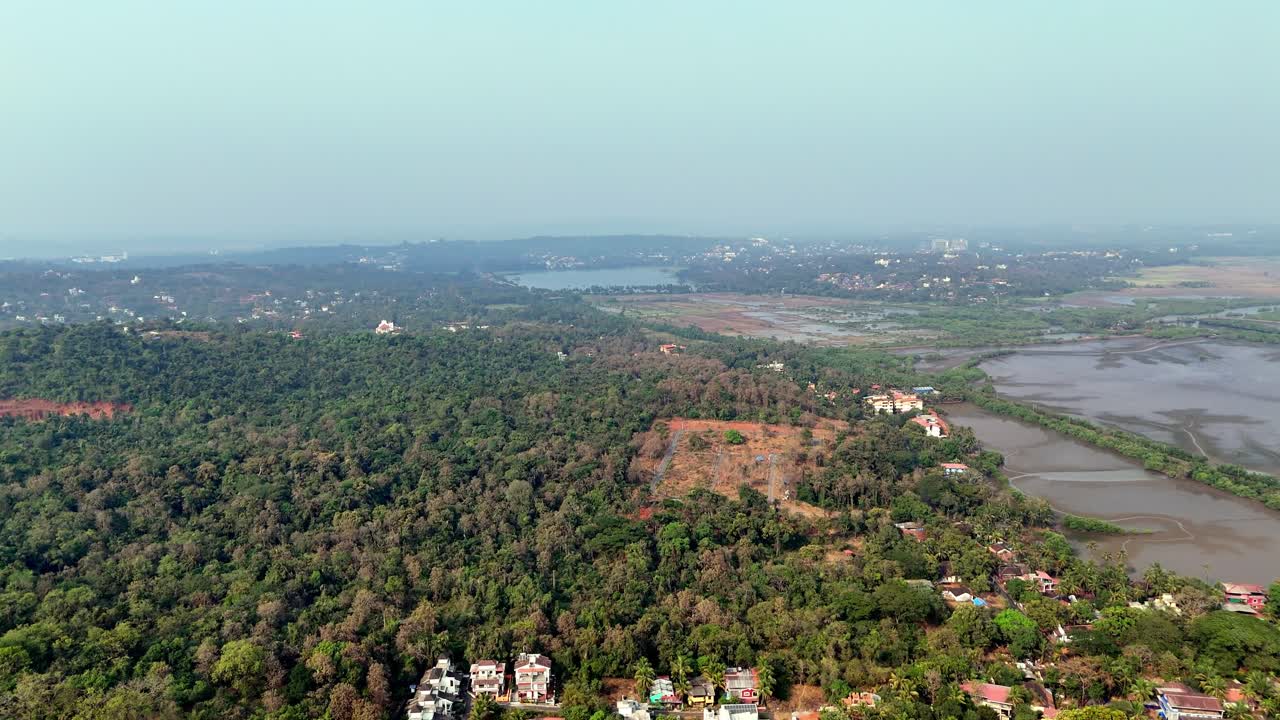 Veduta aerea cinematografica di Ribandar che mostra una fitta copertura forestale verde che si estende verso fiumi e zone umide, con case sparse e un paesaggio urbano distante sotto un cielo sereno