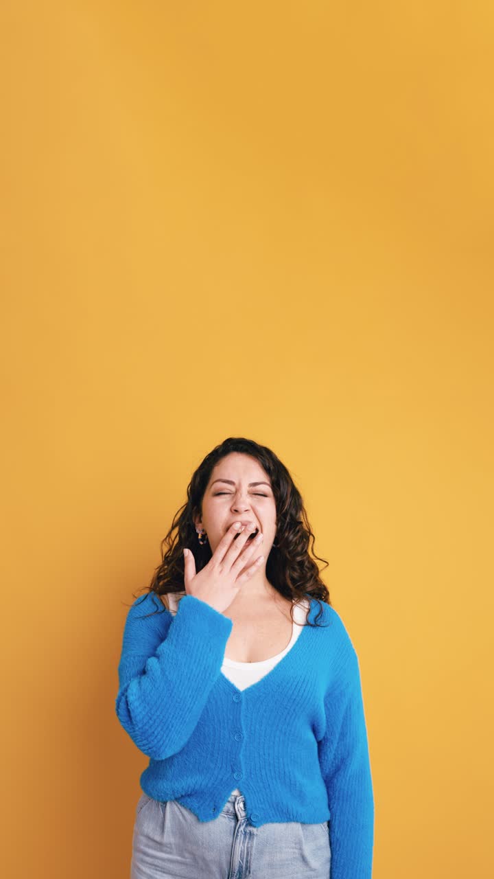 Giovane donna che sbadiglia e si copre la bocca su sfondo giallo.