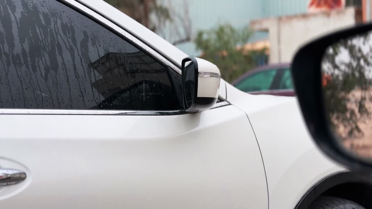 Primo piano dello specchietto laterale di un'auto bianca e di un finestrino striato di pioggia, parcheggiata all'aperto vicino a edifici, catturato in una luce diurna soffusa con profondità di campo ridotta e un'atmosfera urbana umida.