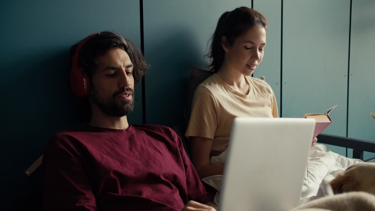 A brunette guy in a red t-shirt wireless headphones communicates via video communication at home. Near the guy sits his girlfriend and reads a book. Happy couple at home