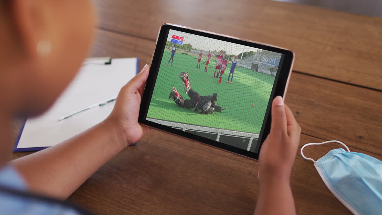 태블릿에서 하키 경기를 시청하는 얼굴 마스크와 함께 테이블에 앉아있는 여성의 복합