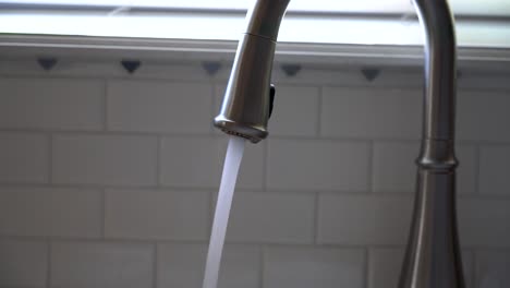 a close shot as water runs from a curved stainless steel kitchen faucet in front of a white tile backsplash with blue diamond shapes, beneath a kitchen window.