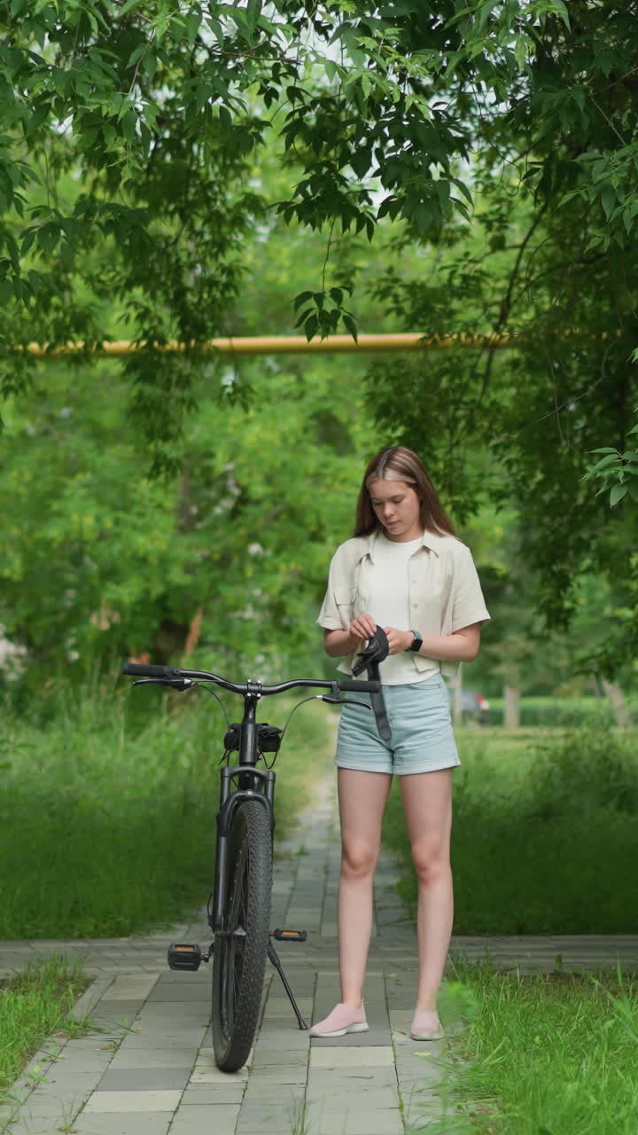 검은 장갑을 들고 있는 여성이 포장된 길에서 주차된 자전거에 접근하고, 오른손에 스마트워치를 착용하면서 장갑을 착용할 준비를 하고, 배경에는 울창한 초록색, 나무 및 주거 건물이 포함되어 있습니다.