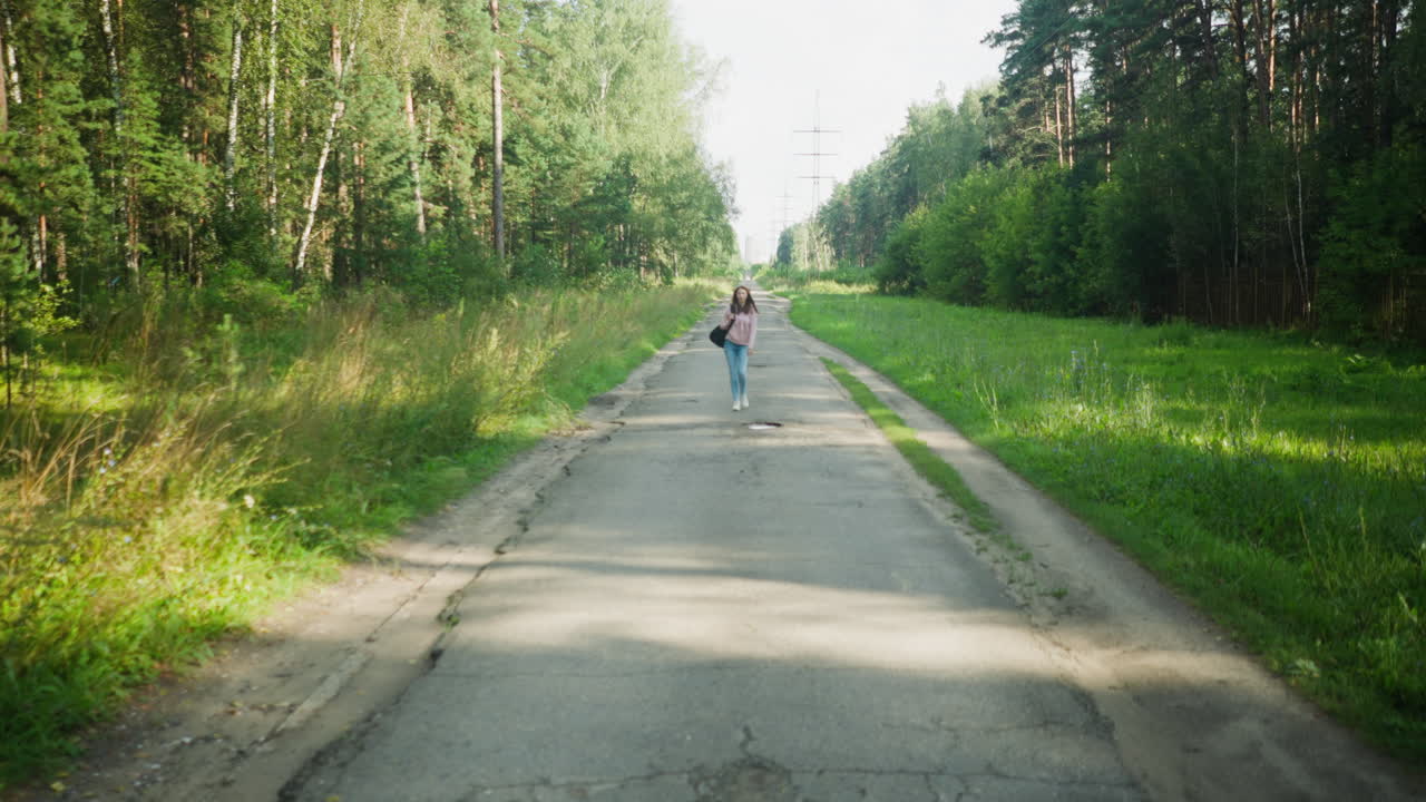 Young woman in casual pink hoodie and jeans walking alone down long empty road surrounded by forest, carrying shoulder bag and heading home in daylight, casting long shadow on cracked pavement