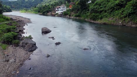 Aerial-dolly-along-slow-moving-river-in-lush-green-jungle-with-luxury-home-on-cliffside